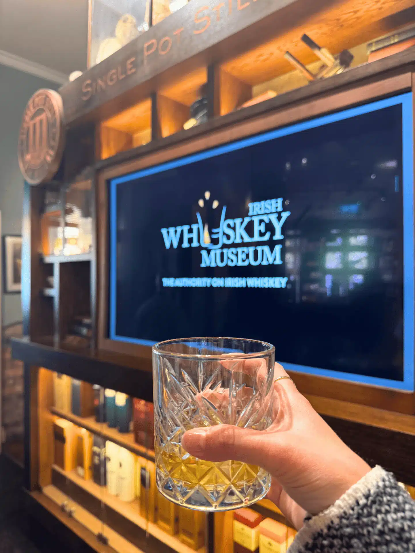 Home 32 A hand holds up an empty crystal-cut whiskey glass in front of a glowing screen displaying the Irish Whiskey Museum logo and the tagline The Authority on Irish Whiskey, framed by a Single Pot Still display shelf inside the museum in Dublin, Ireland.