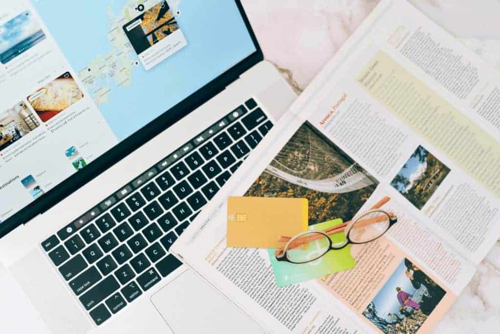 A laptop open with a map from a booking platform, and a magazine open with glasses, credit cards, and a post-it note on top.