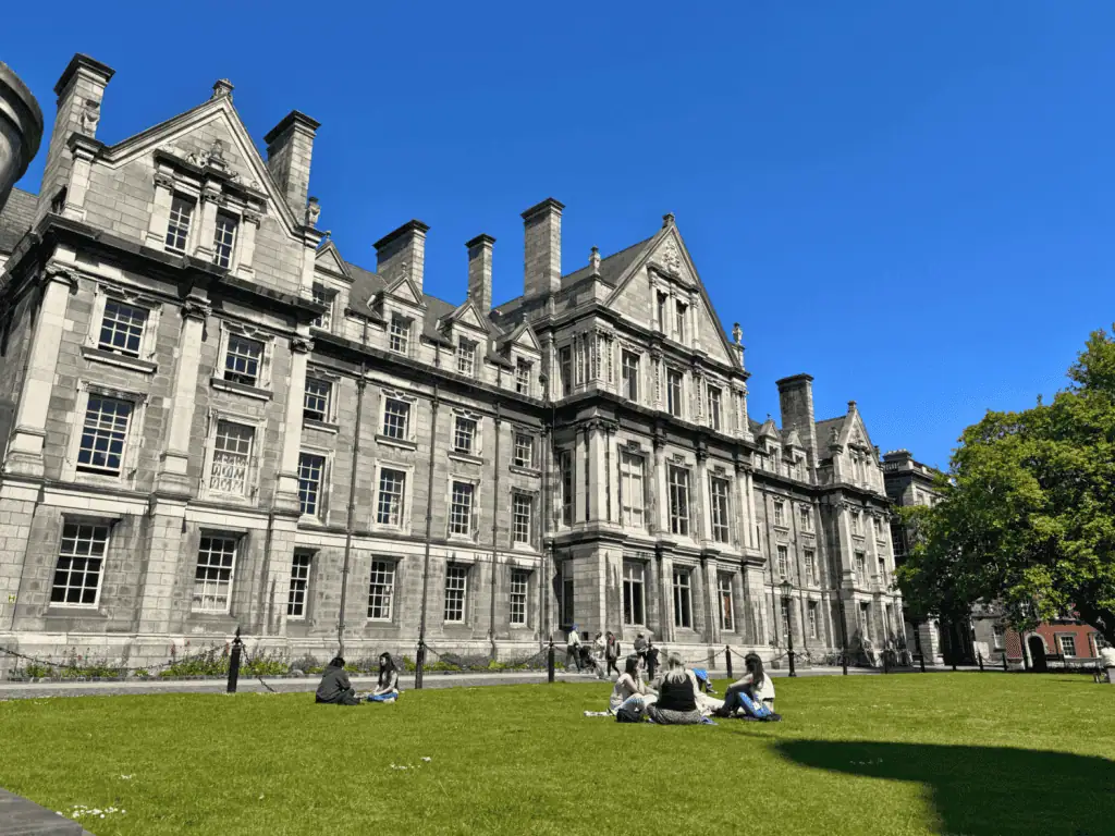 2-Day Dublin, Ireland Itinerary: Top Things to Do for First-Timers 9 Small groups of students sit and chat on a sunlit green lawn in front of an ornate gray granite Victorian building with steep gabled rooflines and tall brick chimneys in the historic front square of Trinity College Dublin, in Dublin, Ireland, under a clear blue sky.