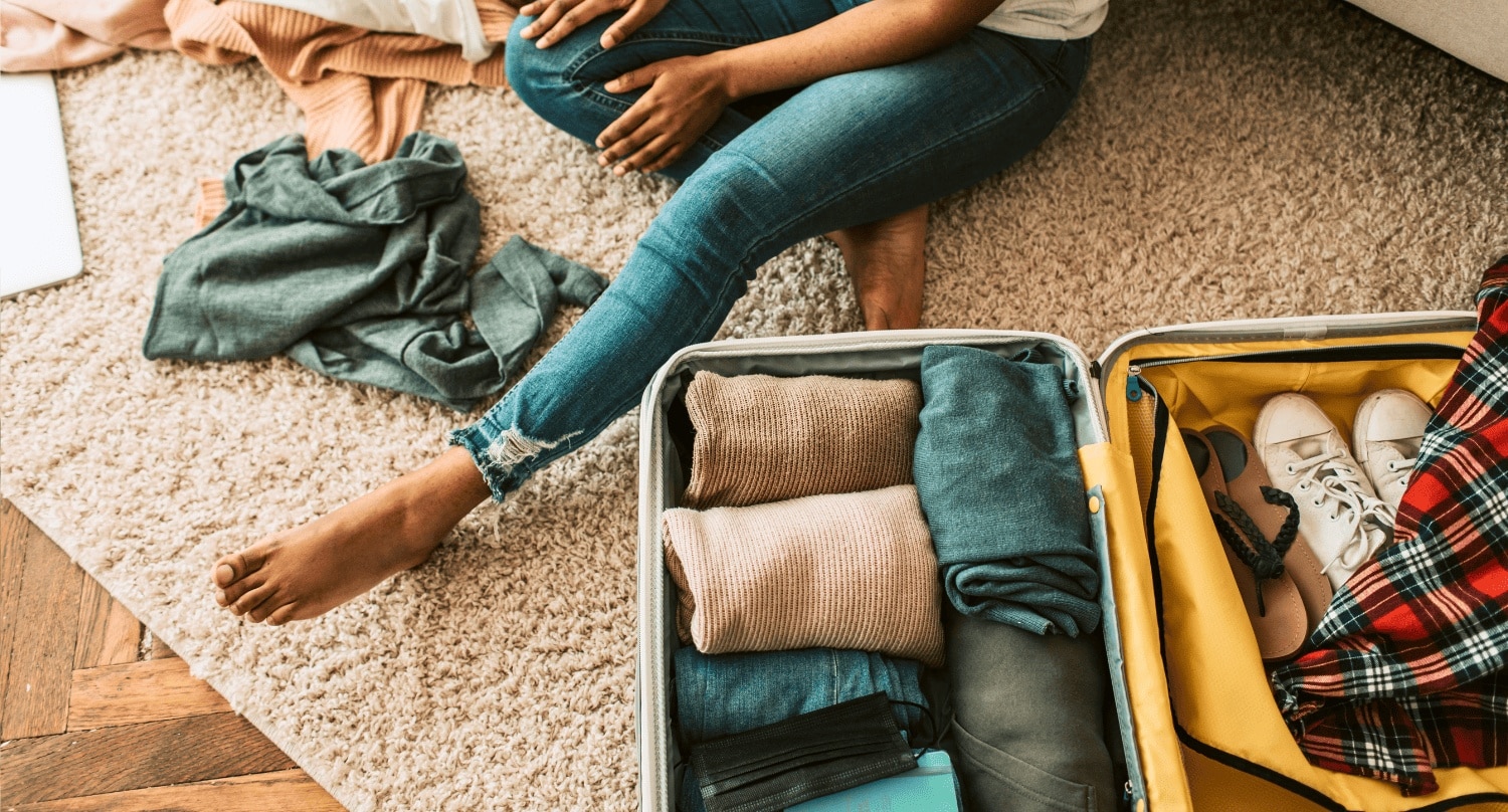 Woman seated on a carpeted rug packing a suitcase with clothes and shoes.