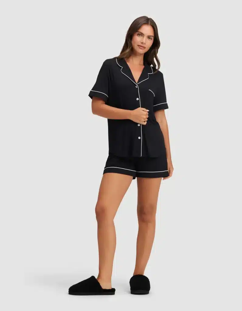 What to Wear in Greece: A Practical Packing List for Women 11 Woman with long brown hair models a black women's Cozy Earth bamboo stretch knit classic pajama short set featuring a button-front short-sleeve top with notched collar, white contrast piping trim, chest pocket, and matching black shorts with white piped hem, paired with black fluffy slip-on slippers, against a light gray background.