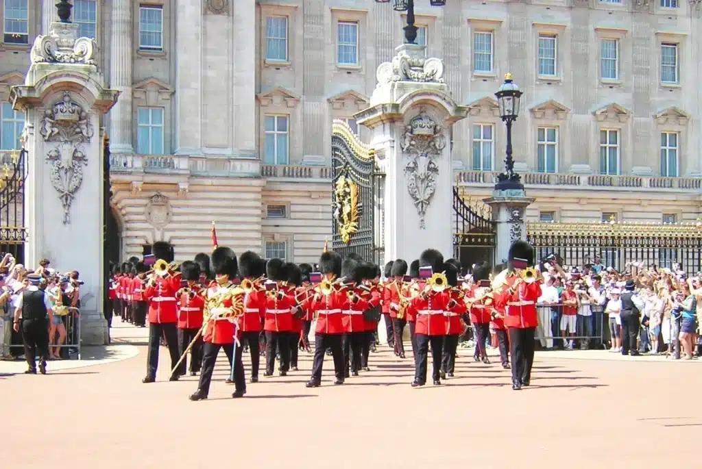 London 2-Day Itinerary: The Best Attractions for First-Timers + Downloadable Map 10 Guards in red tunics and bearskin hats marching in formation in front of Buckingham Palace, London, England, United Kingdom, during the Changing of the Guard ceremony, with the palace gates and a crowd of onlookers in the background.