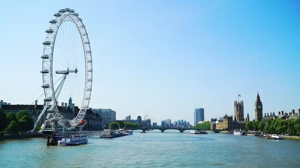 London 2-Day Itinerary: The Best Attractions for First-Timers + Downloadable Map 35 The London Eye observation wheel rises tall beside the Thames River, with tour boats and barges passing below and the Houses of Parliament and the clock tower of Big Ben visible in the distance under a clear blue sky in central London, England, United Kingdom.