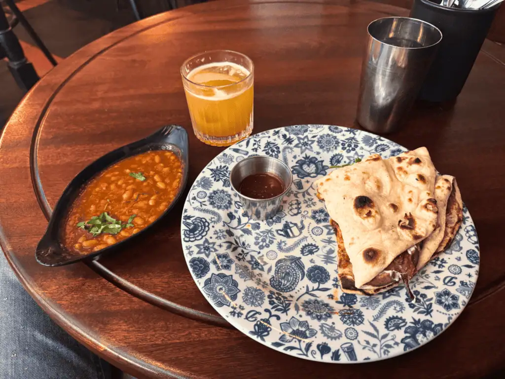 London 2-Day Itinerary: The Best Attractions for First-Timers + Downloadable Map 5 Dishoom Naan Roll with Tomato-chilli jam, a side of beans, and an orange cocktail on a wooden table served in London, England, United Kingdom.