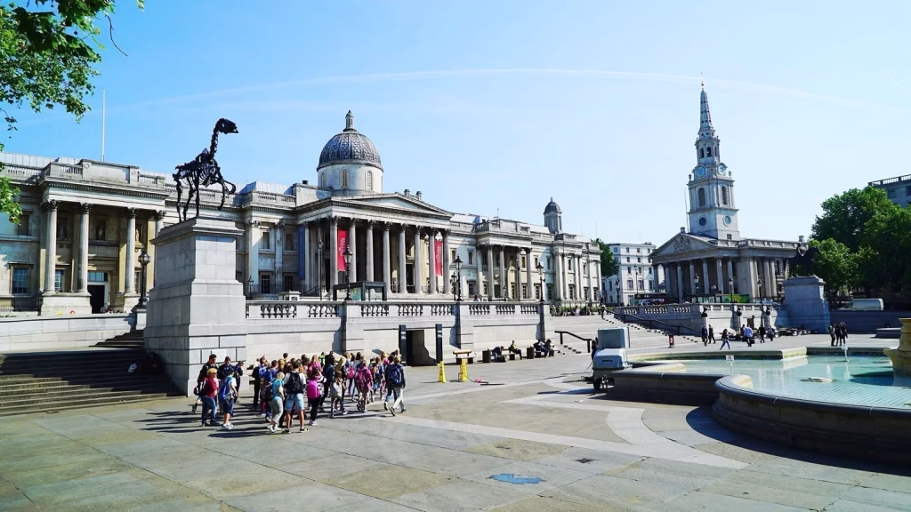Film & TV Tourism 22 Trafalgar Square in London, England, United Kingdom, with the grand neoclassical facade of the National Gallery prominently in the background. To the right, the spire of St Martin-in-the-Fields church rises above the surrounding buildings, while people gather around the square’s fountains and monuments in the foreground.