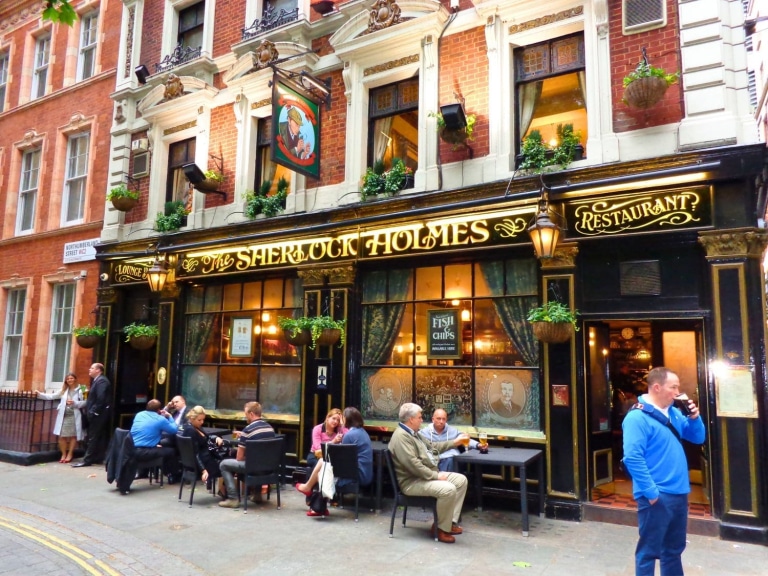 Sherlock Holmes London Walking Tour: Free Self-Guided Literary Route + Downloadable Map 15 Diners and drinkers relax at outdoor tables beneath hanging flower baskets and a painted sign depicting Sherlock Holmes outside the black-and-gold Victorian facade of The Sherlock Holmes pub on Northumberland Street, near Trafalgar Square in central London, England, United Kingdom.