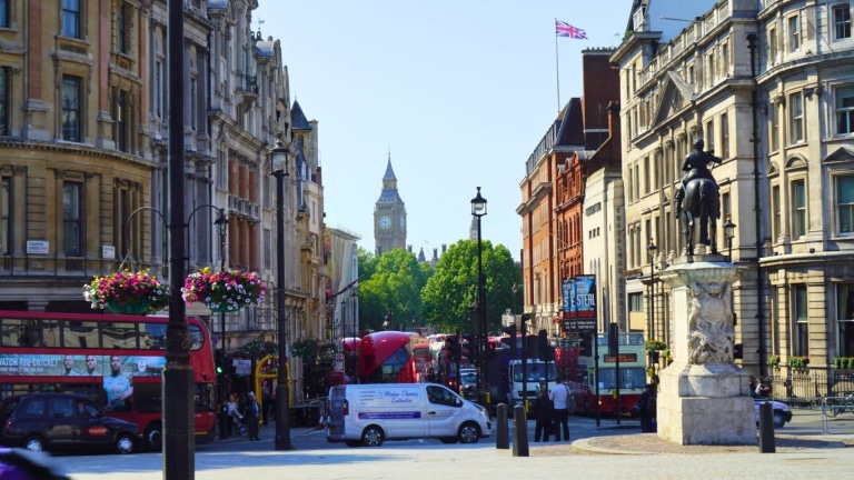 Red double-decker buses and black cabs fill a busy street lined with historic stone buildings, colorful hanging flower baskets, and a bronze equestrian statue, leading toward the distant clock tower of Big Ben near Trafalgar Square in central London, England, United Kingdom.