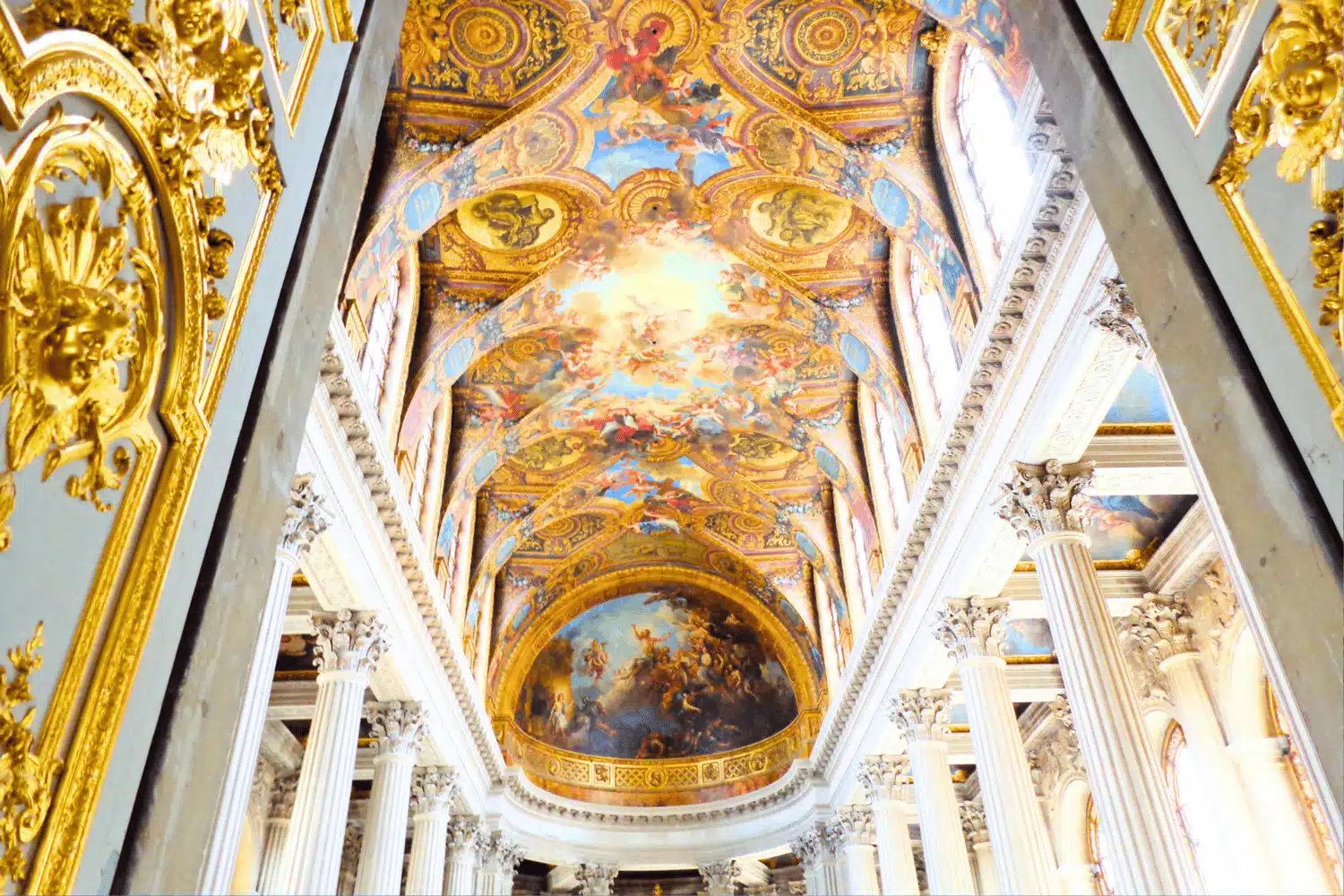 Ornate ceiling in the Royal Chapel at the Palace of Versailles in Versailles, France, richly decorated with gold trim and classical frescoes depicting mythological scenes lined on either side by white columns.