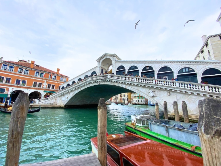 Rialto Bridge arches over the Grand Canal in Venice, Italy, with gondolas and boats gliding through the water below. Historic buildings line the canal, capturing the charm and architecture of Venice.