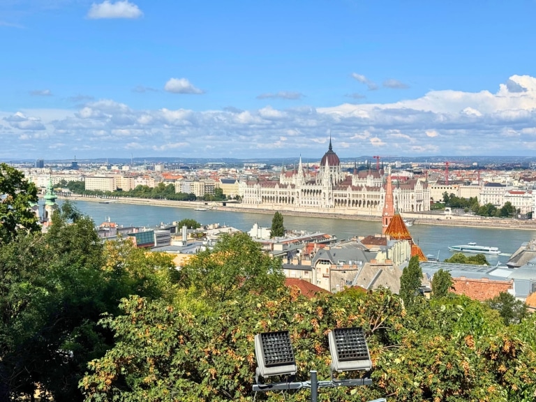A panoramic view of Budapest, Hungary, with the iconic Hungarian Parliament Building prominently displayed along the Danube River under a bright blue sky. In the foreground, leafy trees and rooftops lead the eye toward the neo-Gothic architecture and the riverboat cruising on the water.