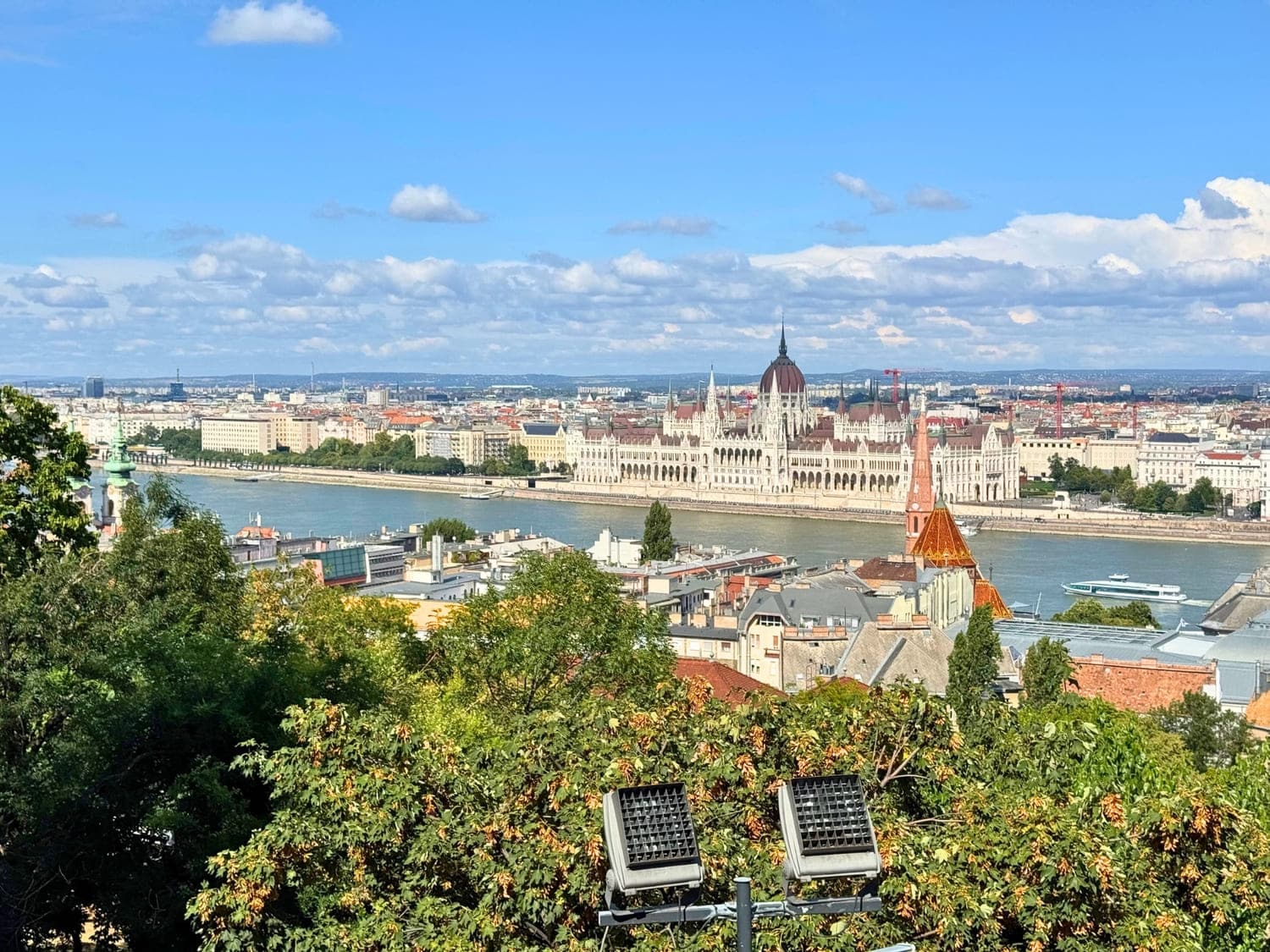 A panoramic view of Budapest, Hungary, with the iconic Hungarian Parliament Building prominently displayed along the Danube River under a bright blue sky. In the foreground, leafy trees and rooftops lead the eye toward the neo-Gothic architecture and the riverboat cruising on the water.