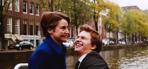 75 Movies Set in European Destinations to Inspire Your Next Trip 72 Animated GIF Scene from The Fault in Our Stars showing Hazel and Gus laughing together on a boat ride through an Amsterdam canal. Hazel wears a nasal cannula and a blue coat while Gus leans in smiling, surrounded by tree-lined streets and classic Dutch buildings.