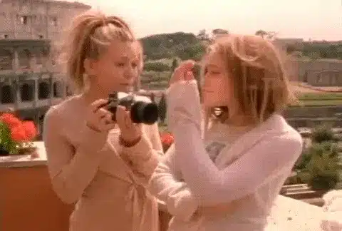 75 Movies Set in European Destinations to Inspire Your Next Trip 66 Animated GIF from When in Rome (2001) showing Mary-Kate and Ashley Olsen on a scenic balcony with the Colosseum in the background. One twin holds a camera while the other adjusts her hair.