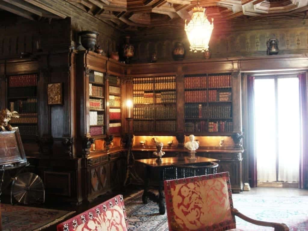 Hearst Castle Tours: Which is Best for First-Timers + An Honest Review 12 The lavish library at Hearst Castle in San Simeon, California, USA, featuring rich wood‑paneled bookshelves, a Gothic‑inspired vaulted ceiling, ornate lighting fixture, a table with busts on it, and a plush seating area.