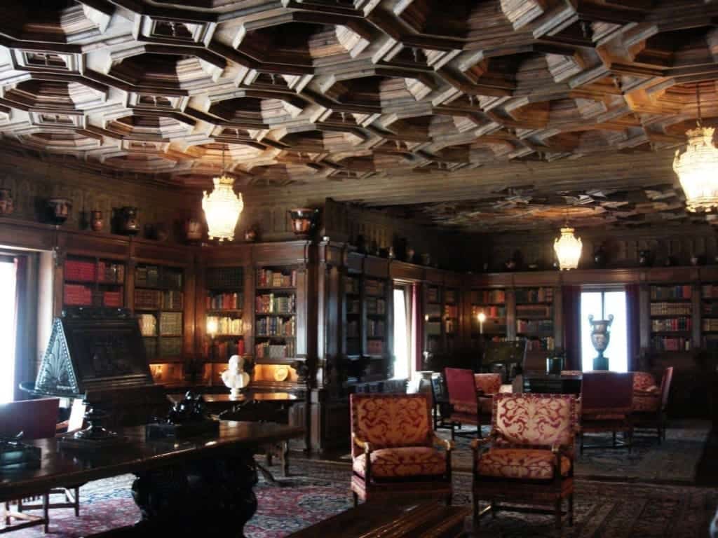 Hearst Castle Tours: Which is Best for First-Timers + An Honest Review 11 The Hearst Castle Library is filled with shelves of books, upholstered and patterned chairs, chandeliers, and an ornate wooden ceiling as sunlight pours in at the San Simeon, California, USA, estate.