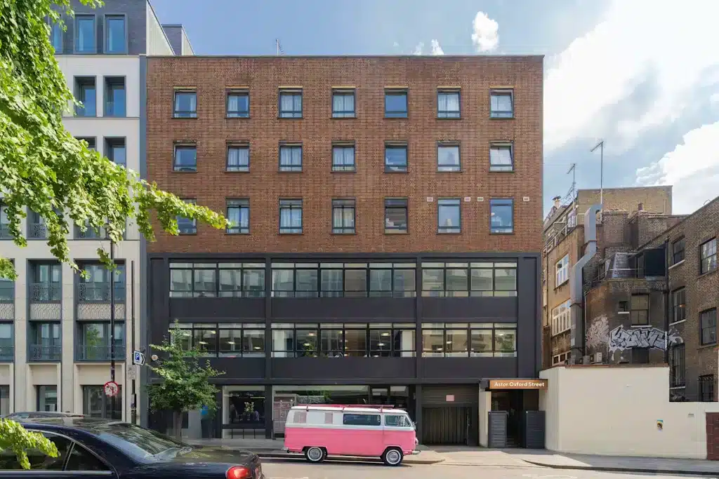 Reviewing Astor Oxford Street: Is It a Safe Hostel to Stay at in Soho, London? 4 Street view of a modern, boxy brick and black-trimmed building labeled "Astor Oxford Street" with a vintage pink and white van parked in front. The surrounding area features a mix of older and newer architecture, including a graffiti-covered wall on the right and a leafy tree partially visible on the left in London, England, United Kingdom.