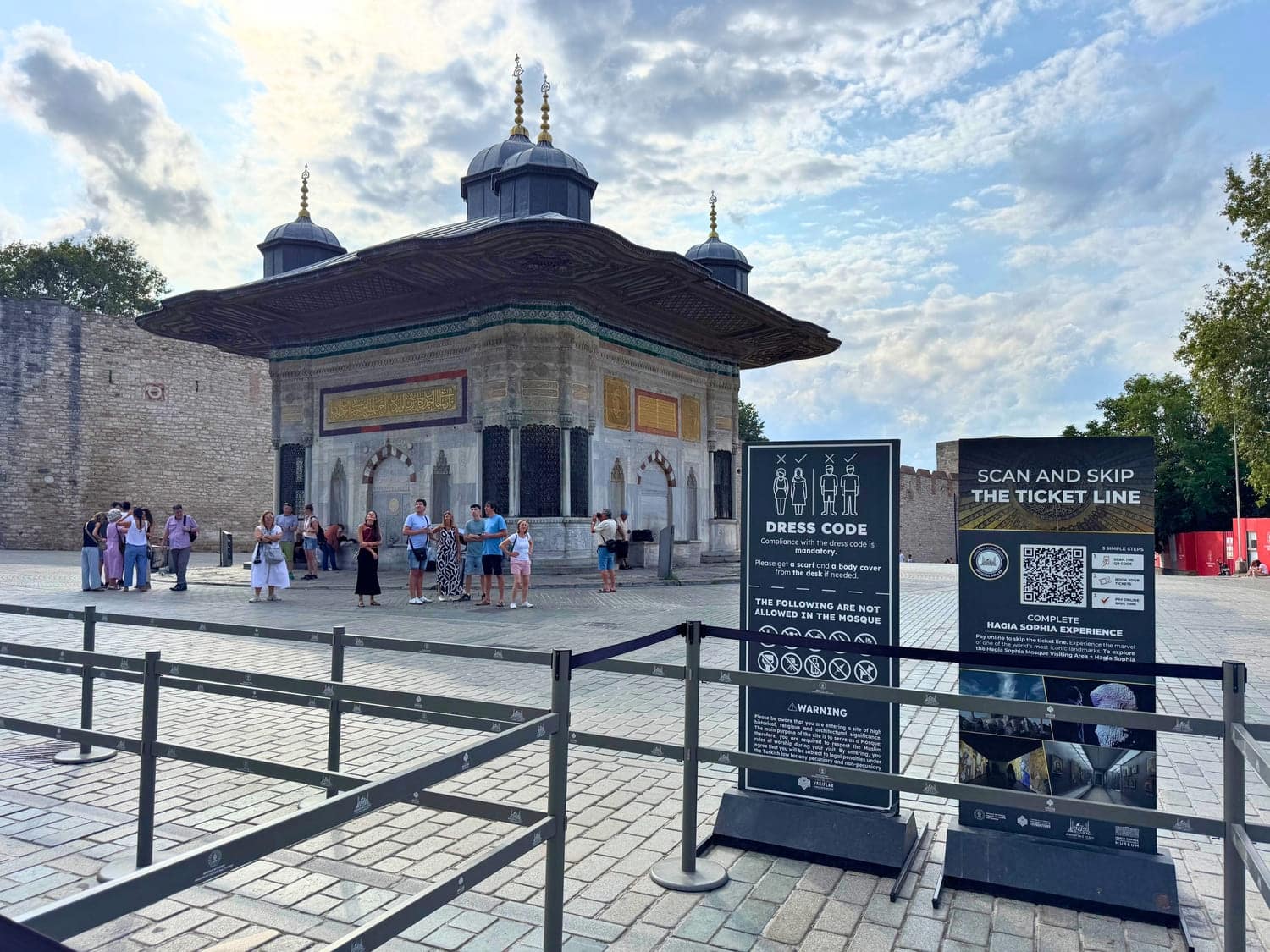 44 Things to Do Before Traveling Internationally + Free Downloadable Checklist 20 Tourists gather near the ornate Fountain of Ahmed III outside Topkapi Palace in Istanbul, Turkey, where two signs are posted: one outlining the mandatory dress code for visiting nearby Hagia Sophia with visual icons, and another promoting a fast-track ticket option with a QR code for the Hagia Sophia experience.
