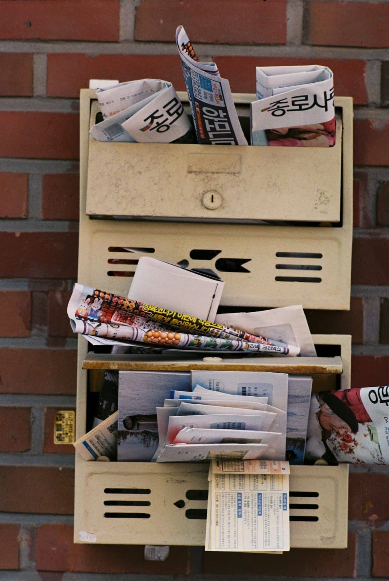 44 Things to Do Before Traveling Internationally + Free Downloadable Checklist 7 Overflowing metal mailbox mounted on a brick wall, filled with rolled newspapers, letters, and printed advertisements in Korean. The mailbox has three compartments, each crammed with mail, including colorful flyers and folded sheets with blue and black text.