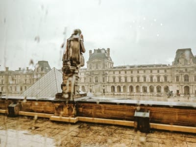 Rain blurring the view from a window at the Louvre Museum in Paris, France, of the wet courtyard with the glass pyramid in the background as overcast skies loom above.