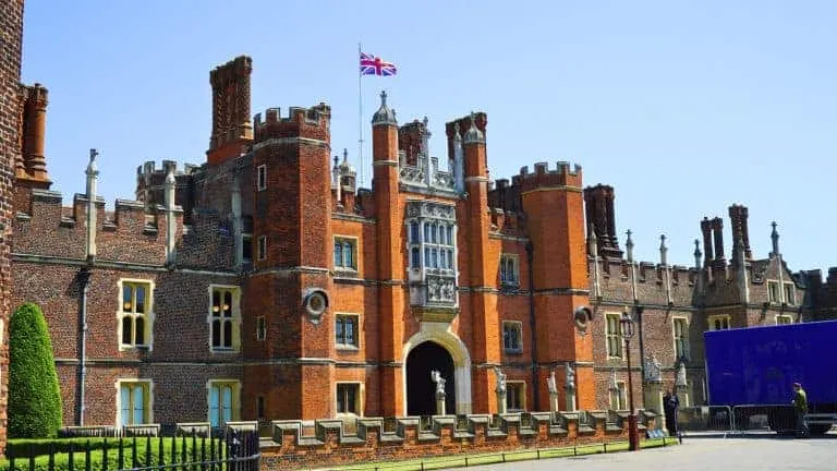 The red‑brick Tudor and Baroque façade of Hampton Court Palace in East Molesey, England, United Kingdom, showcasing tall chimneys, stone mullion windows, and a central arched entrance beneath a clear sky.
