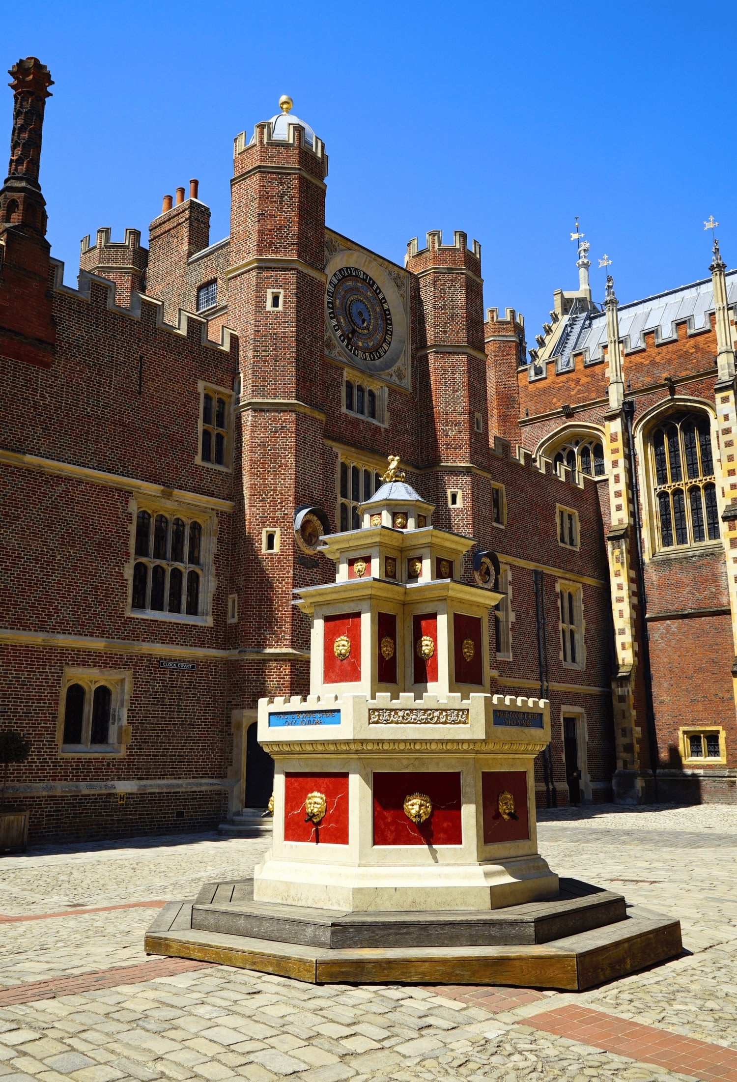 Guide to Visiting Hampton Court Palace on a Short Day Trip from London 3 Ornate Tudor‑style wine fountain standing in the Base Court of Hampton Court Palace in East Molesey, England, United Kingdom, featuring red and gold panels, gold‑lion spouts and a central tiered structure of stone and timber with the Astronomical Clock above.