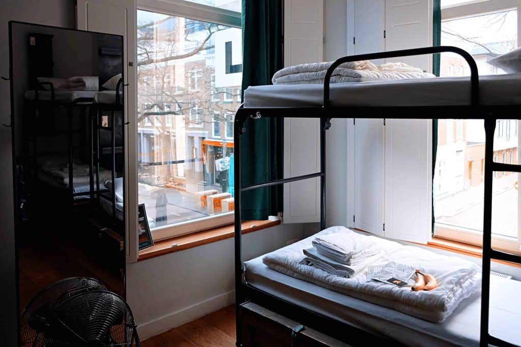 My Experience Staying in a Hostel as a Solo Female Over 35: Honest Insights 9 Hostel room with metal bunk beds, white bedding, a mirror, and lockers in the corner with a window looking out onto the street.