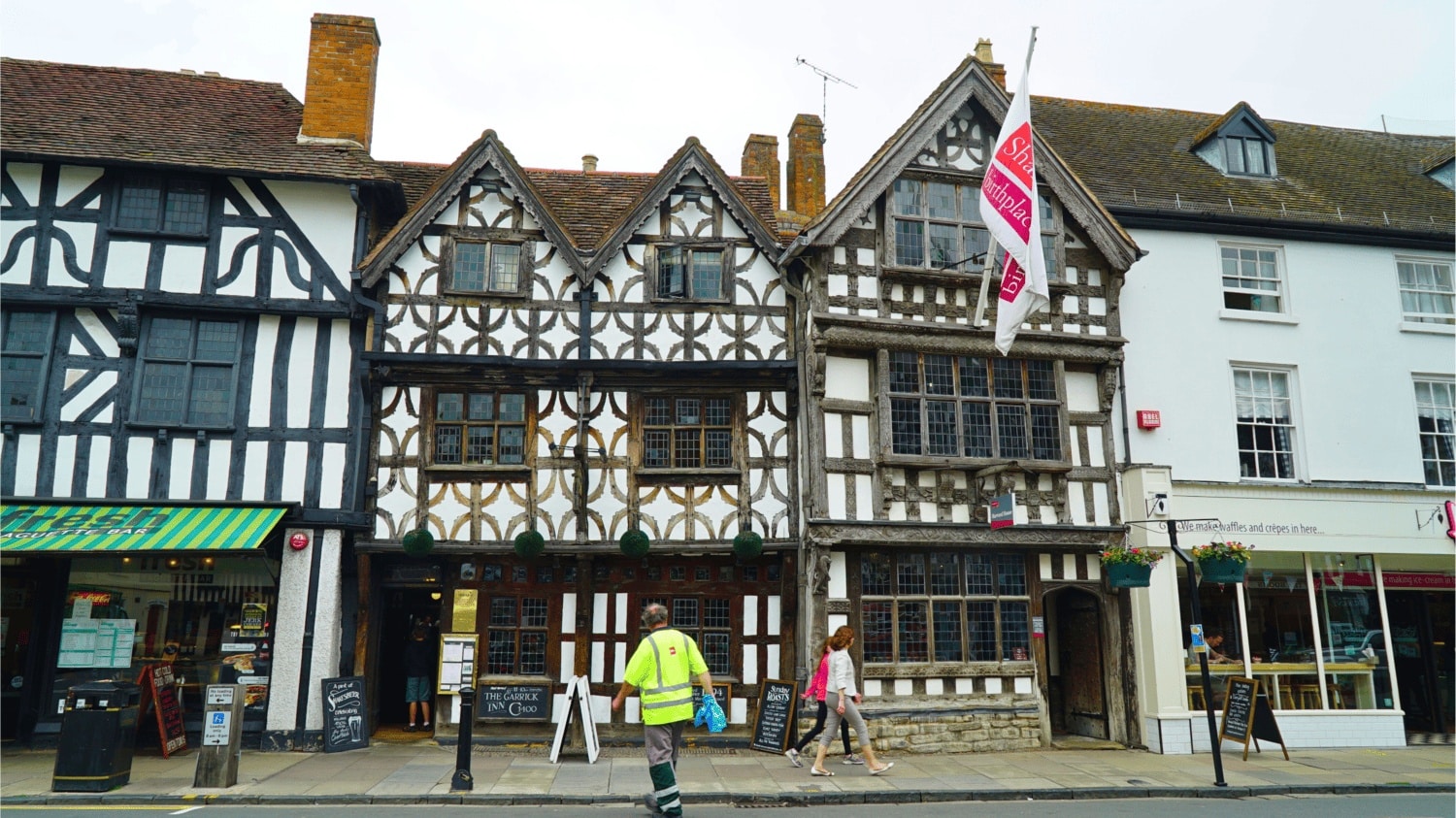 Harvard House, a Tudor-style building with timber framing and a Shakespeare Birthplace Trust flag flying above the entrance, with visitors passing by on the street, in Stratford-upon-Avon, Warwickshire, England, United Kingdom.