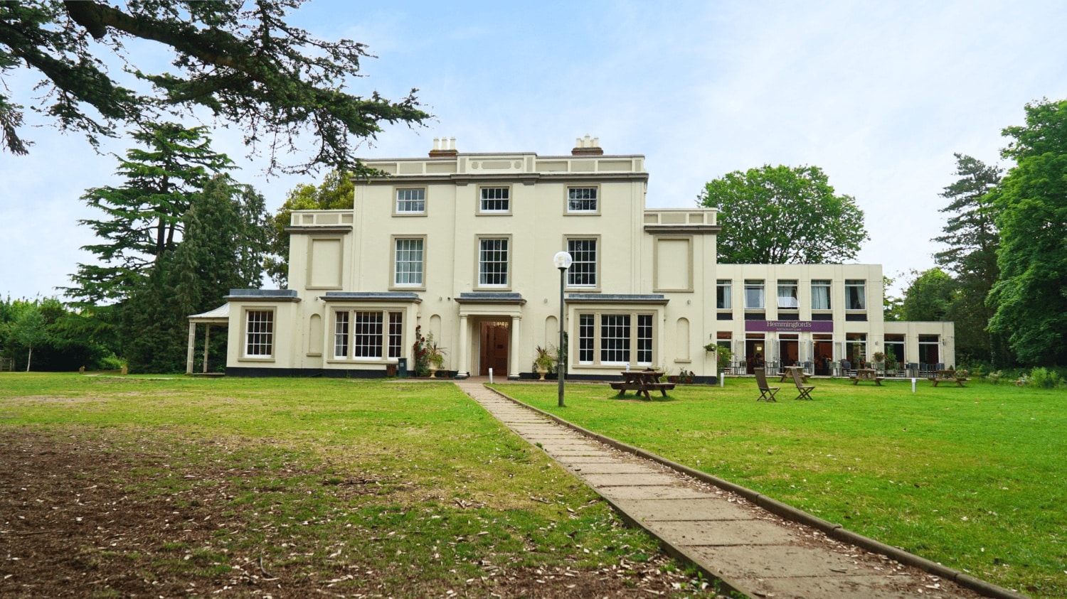 The elegant pale‑cream Georgian façade of YHA Stratford Upon Avon, set within greenery and showing tall sash windows and a central doorway beneath a clear sky. The exterior hints at a stately hostel experience nestled just outside the heart of Stratford‑upon‑Avon, England, United Kingdom.