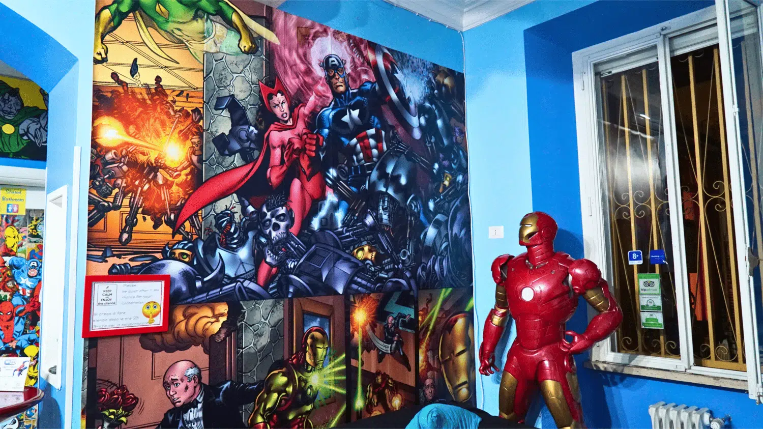 Vibrant wall mural of Marvel comic book characters and a statue of Iron Man inside the themed common area of Comics Guesthouse, a hostel in Rome, Italy, with brightly colored décor and gaming lounge features.