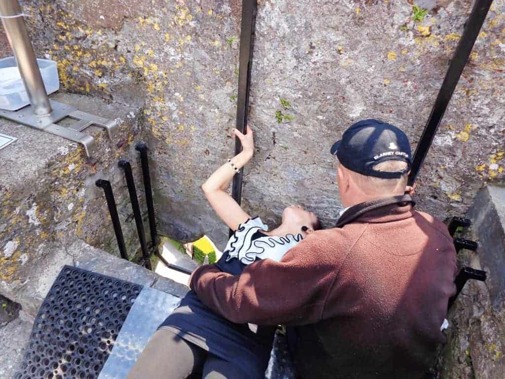 Day Trips from Dublin Without a Car: 6 Easy Irish Escapes by Train or Tour Bus 18 Tourist leaning backward to kiss the Blarney Stone at Blarney Castle in Ireland, supported by a metal railing and guide, with stone walls and greenery visible around the historic site.