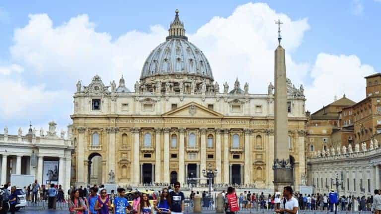 Exterior view of St. Peter’s Basilica in Vatican City with its grand dome rising above the ornate façade. The plaza in front is filled with tourists and features a tall obelisk under a clear blue sky.