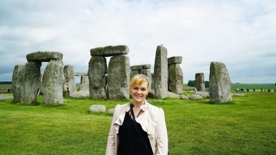 The ancient stone circle of Stonehenge on Salisbury Plain in England, United Kingdom, with Michelle L. Jensen (Travel HerStory) smiling in front of weathered upright megaliths arranged in a ring beneath a partly cloudy sky.