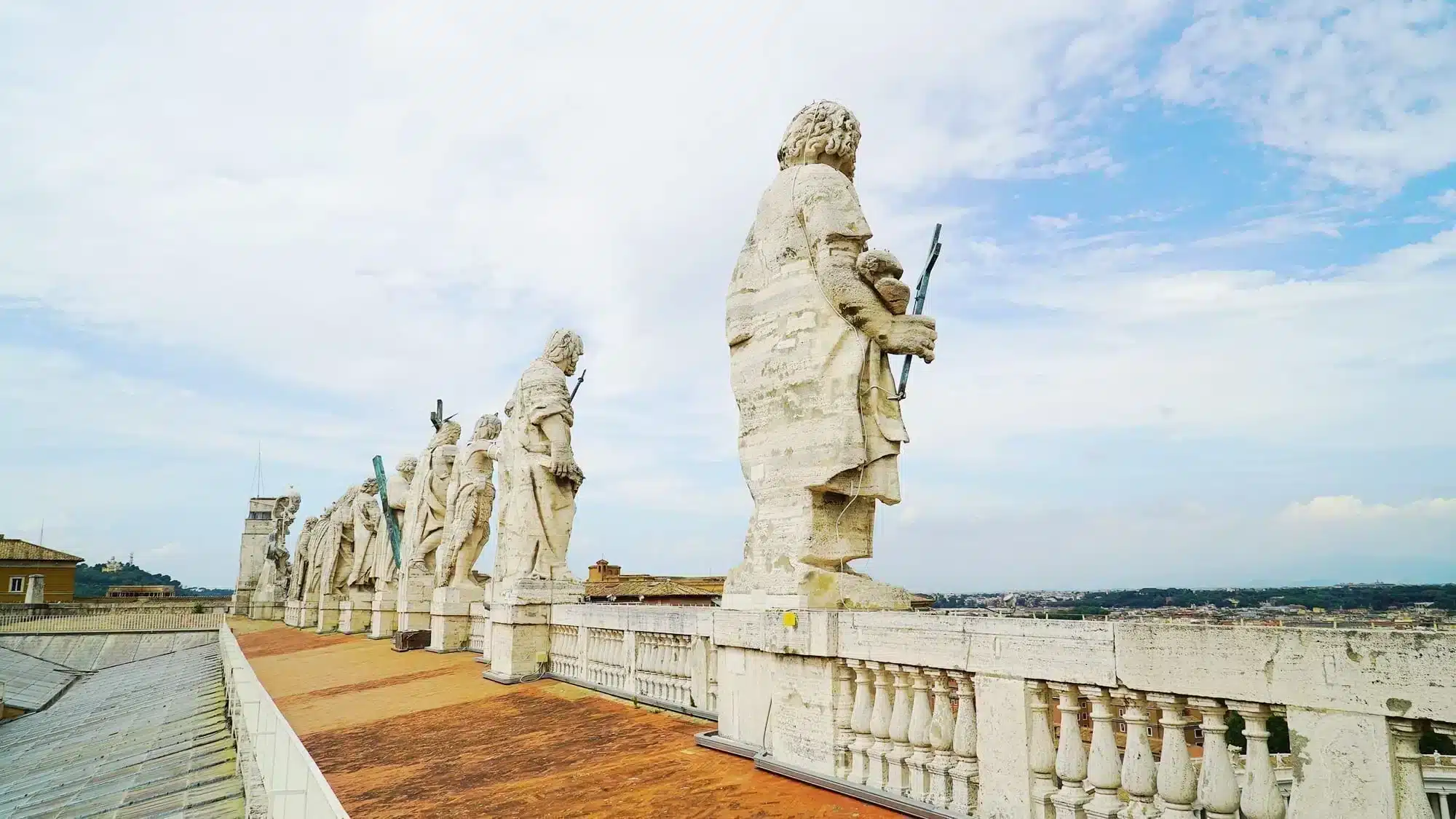 How to Climb St. Peter's Dome + The Mistakes to Avoid That I Made 20 Row of weathered travertine apostle statues lining the lower rooftop terrace of St. Peter's Basilica, Vatican City, each holding a cross or religious symbol, with a sweeping panoramic view of Rome's rooftops and hills stretching to the horizon under a partly cloudy sky.