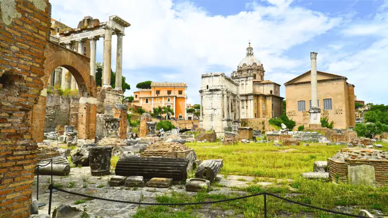 Panoramic view of the Roman Forum and Palatine Hill with ancient ruins, columns, and greenery under a blue sky in Rome, Italy.