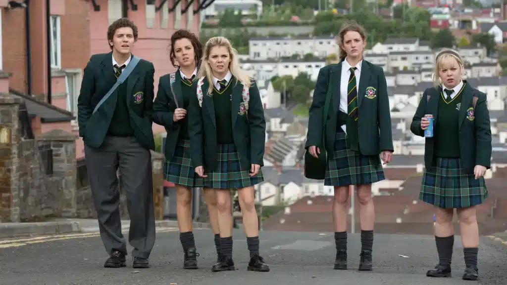 24 TV Series Set in Europe That'll Inspire Your Next Vacation 18 Promotional still from Derry Girls showing five teenagers in green school blazers and tartan plaid skirts walking down a steep Northern Ireland street with a densely packed hillside housing estate visible in the background, all wearing expressions of mild alarm
