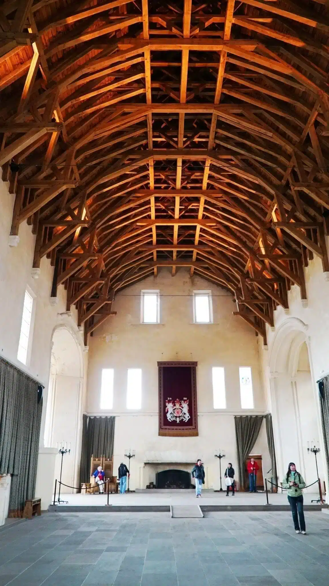 How to Visit Stirling Castle, Scotland: An Easy Day Trip by Train 6 Interior of the Great Hall at Stirling Castle, Scotland, featuring a soaring restored hammer-beam timber roof, whitewashed stone walls, tall narrow windows, a large stone fireplace, and a royal heraldic banner hanging above tourists exploring the medieval banqueting hall.