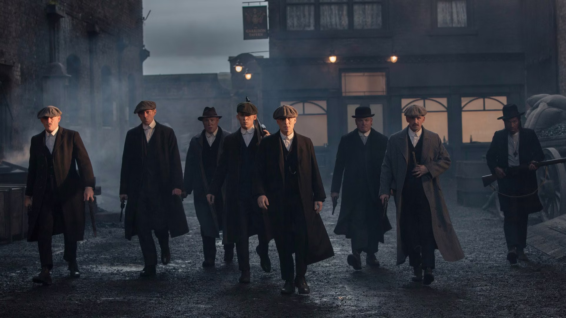 24 TV Series Set in Europe That'll Inspire Your Next Vacation 9 Promotional image from Peaky Blinders showing eight men in 1920s period clothing — flat caps, long overcoats, and suits — walking menacingly through a foggy cobblestone street at dusk, with the Garrison Tavern lit up in the background