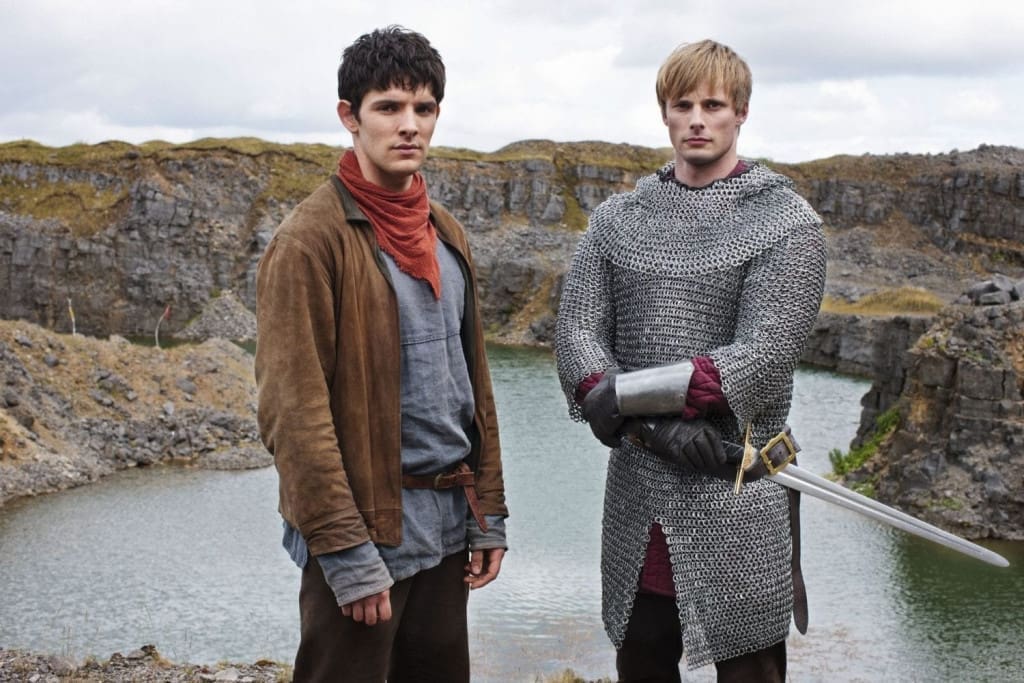 24 TV Series Set in Europe That'll Inspire Your Next Vacation 21 Scene from BBC Merlin TV show, Season 5 Episode 9, showing characters Merlin and Arthur in medieval costumes standing in front of a stone landscape with a water feature.