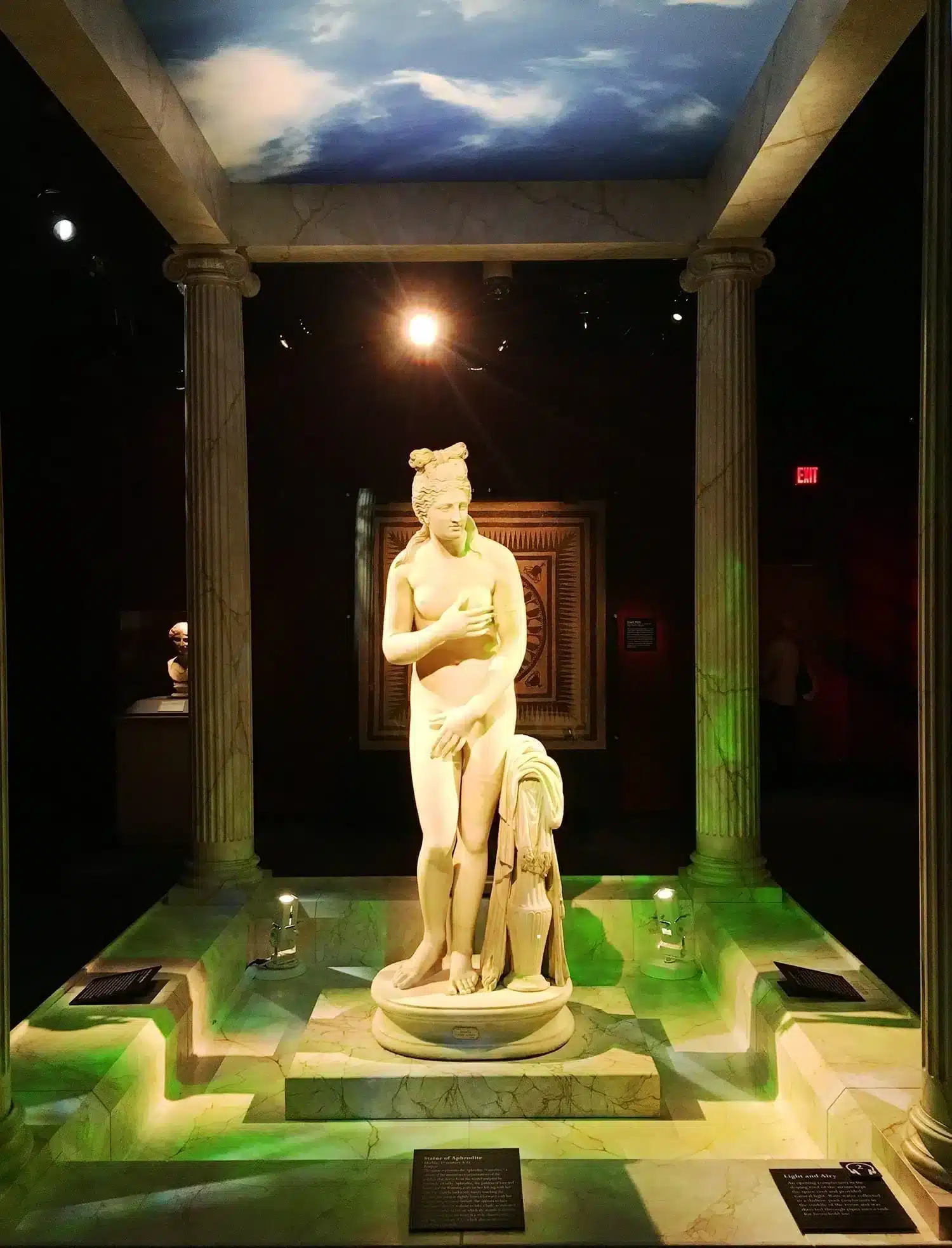 Pompeii: The Exhibition: Guide on What to Expect Before You Visit 10 Marble Statue of Aphrodite displayed in a dramatic Pompeii exhibition gallery with Roman columns, green-lit marble floor, and a painted sky ceiling, spotlit against a dark background with decorative mosaic panel behind.