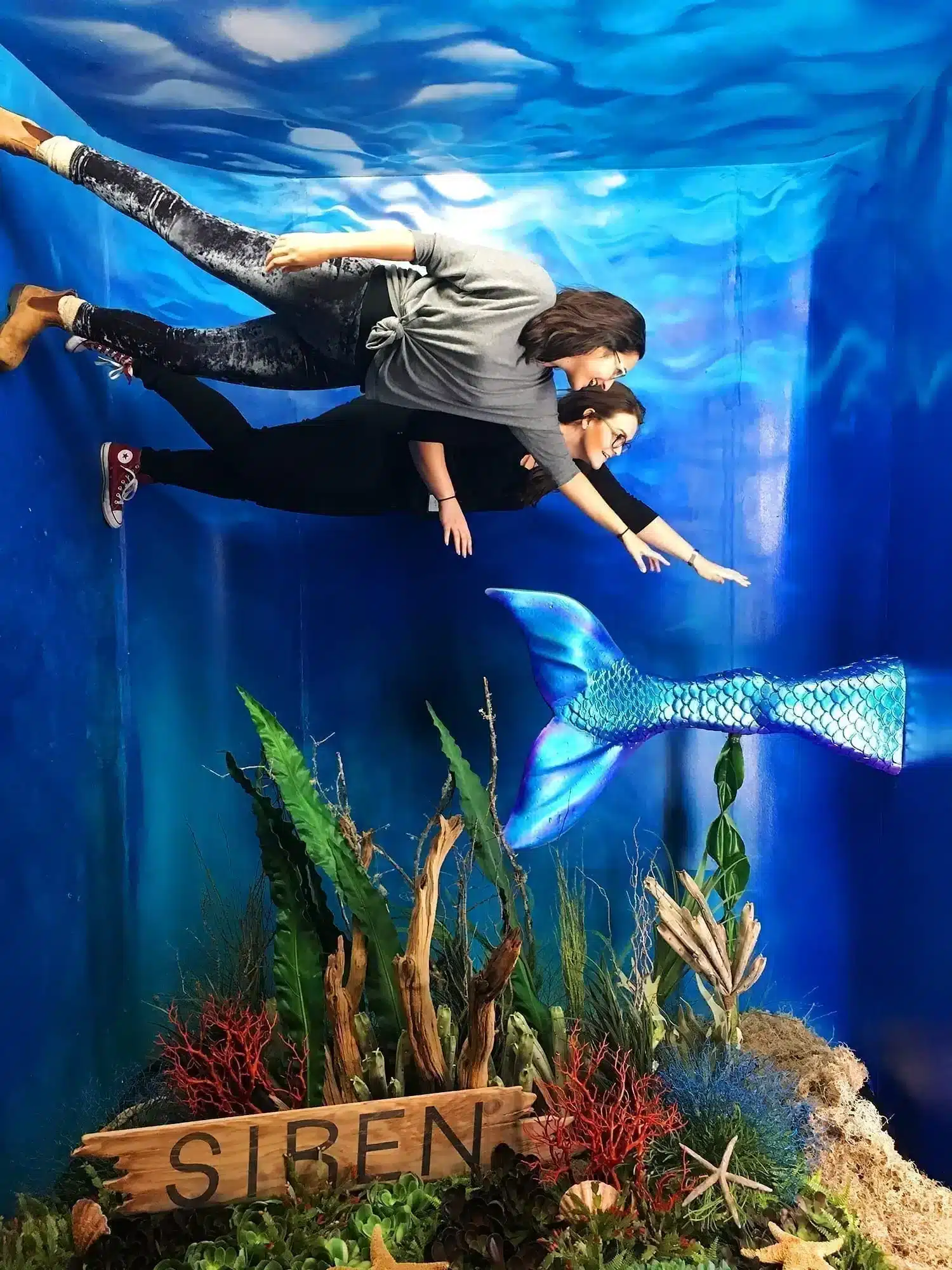21 Cool, Nerdy Things to Do in LA—Activities for Kids to Adults 59 Two women pose in a gravity-defying underwater illusion photo op, appearing to swim alongside a shimmering blue mermaid tail above a seabed of coral and seaweed, with a "Siren" driftwood sign at the base, at the Siren Freeform pop-up mermaid museum.