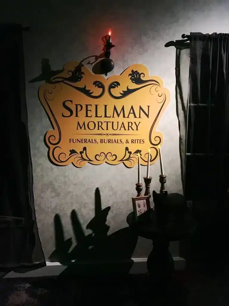 21 Cool, Nerdy Things to Do in LA—Activities for Kids to Adults 54 rnate gold "Spellman Mortuary – Funerals, Burials, & Rites" sign on a dark damask wall, flanked by flickering candles and a wall sconce, at the Chilling Adventures of Sabrina Netflix pop-up experience.
