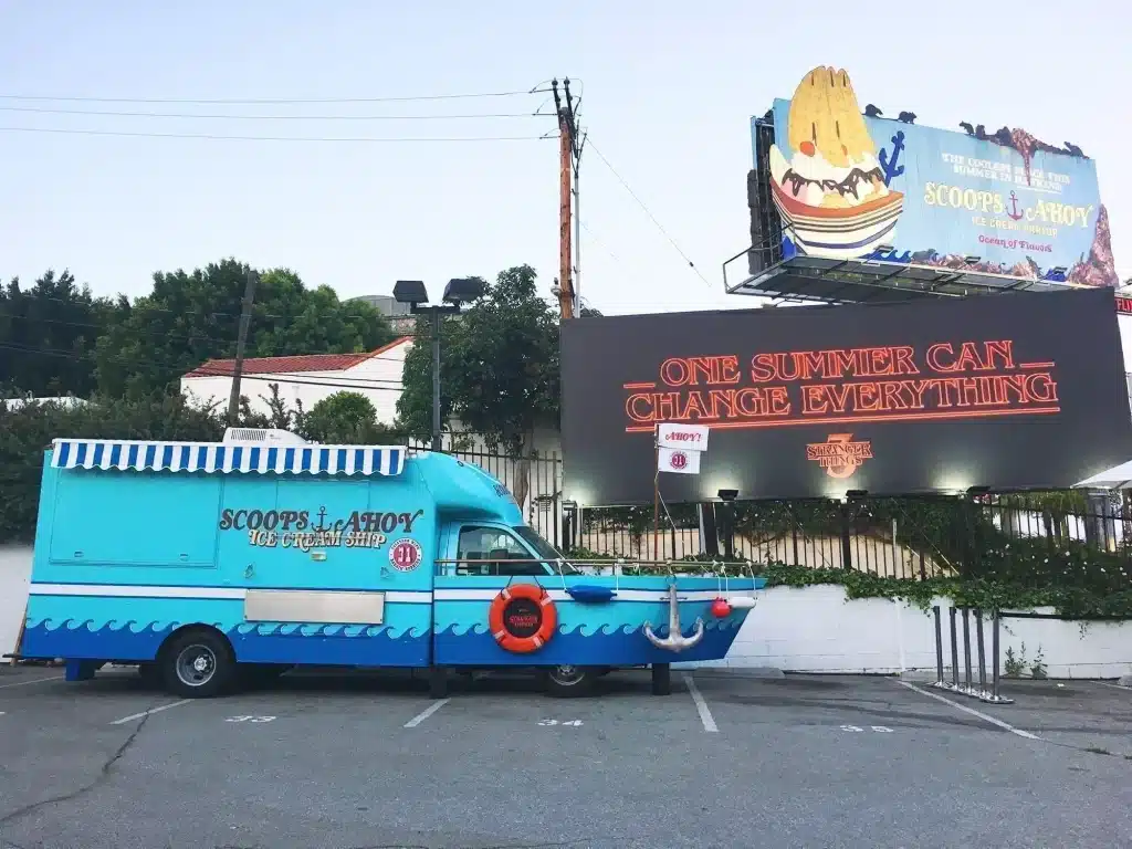21 Cool, Nerdy Things to Do in LA—Activities for Kids to Adults 40 Scoops Ahoy ice cream truck themed as a nautical ship with wave graphics, striped awning, and life preserver ring, parked beside a Stranger Things Season 3 billboard reading "One Summer Can Change Everything" and a Scoops Ahoy Ice Cream Parlor advertisement at the Los Angeles pop-up event.