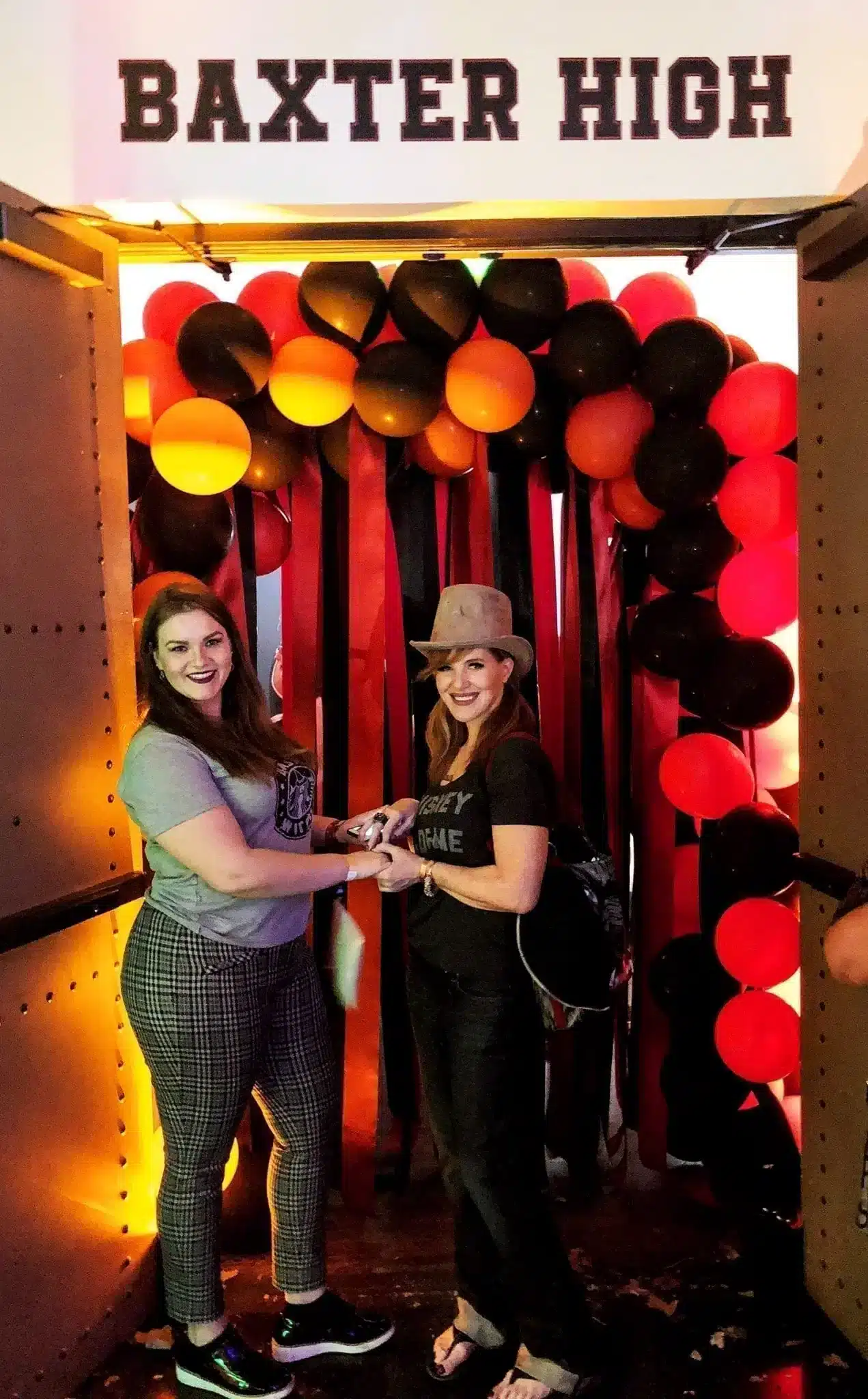 21 Cool, Nerdy Things to Do in LA—Activities for Kids to Adults 56 Two women smiling inside a locker photo op framed by red, orange, and black balloons beneath a "Baxter High" sign at the Chilling Adventures of Sabrina Netflix pop-up event, one wearing a skull hat and the other in plaid trousers.