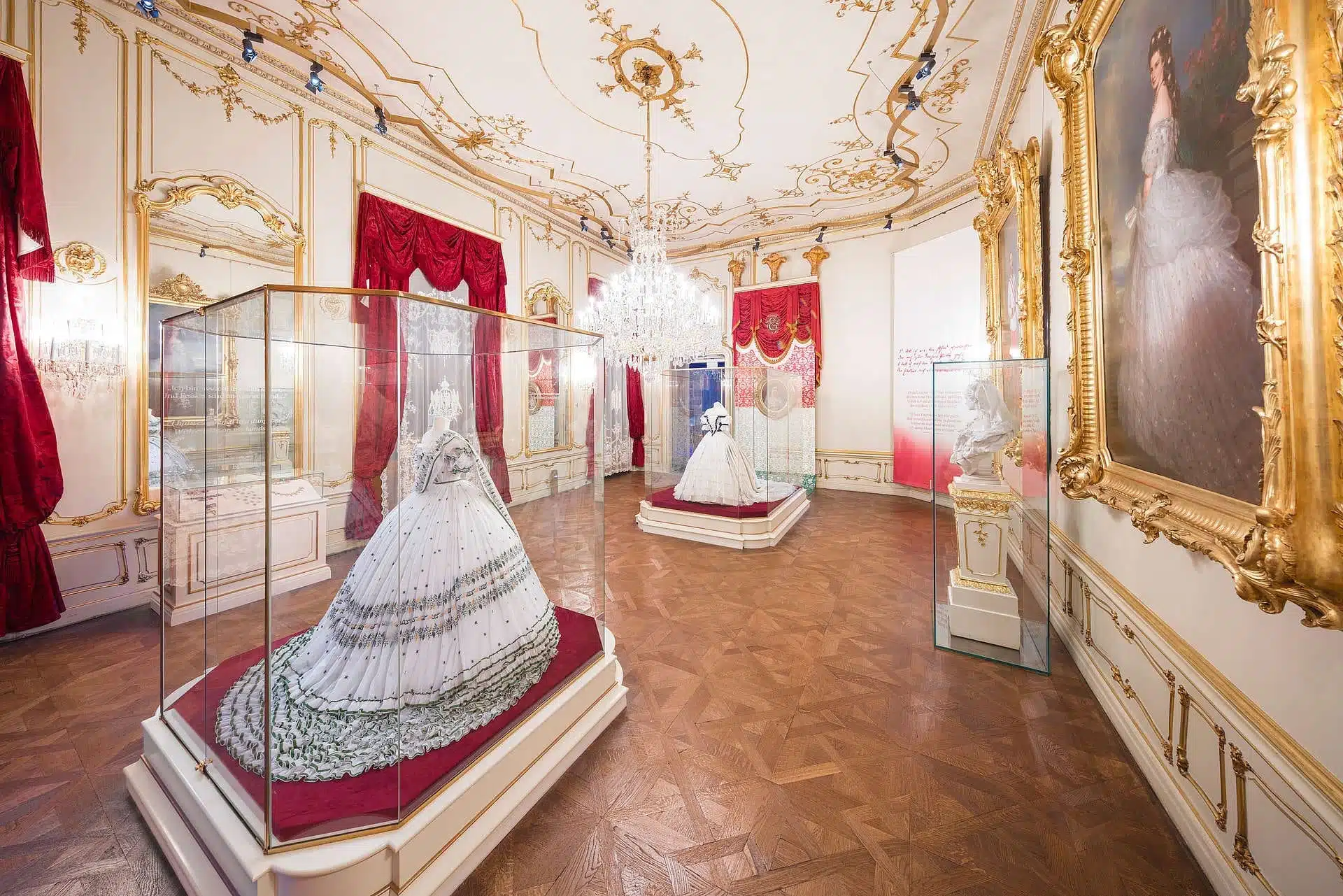 Display of Empress Elisabeth’s gowns at the Sisi Museum in Vienna, Austria, featuring elaborately embroidered 19th-century dresses showcased in glass cases with chandelier lighting and nearby portraits.