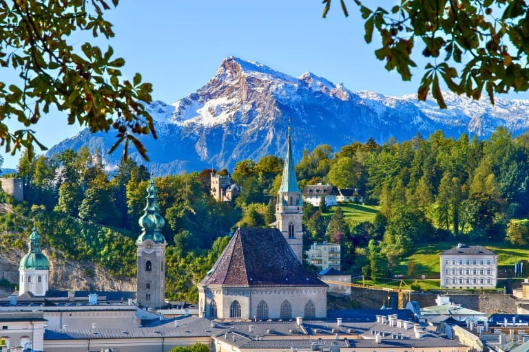 Historic rooftops and church towers amongst greenery in Salzburg, Austria, with snow-capped mountains in the background.
