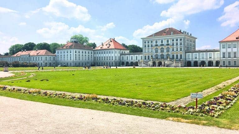 Front view of Nymphenburg Palace in Munich, Germany, with its symmetrical Baroque architecture, as tourists mill around, surrounded by manicured gardens under a blue sky.