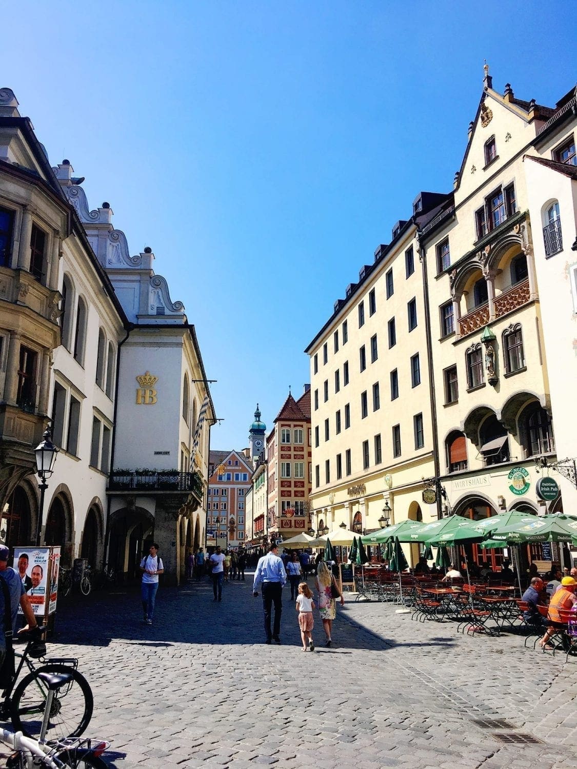 Visiting Old Town Munich, Germany: Comparing Walking Tour & Self-Guided Options 16 Exterior of Hofbräuhaus am Platzl in Munich seen on the left side of a street during a walking tour, featuring traditional Bavarian architecture with arched windows, hanging signs, umbrellas, and groups of people walking by outside.