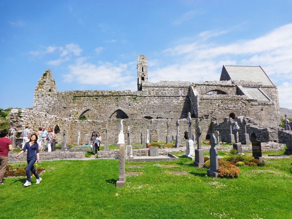 Honest Thoughts on Taking Paddywagon Tours from Dublin to the Cliffs of Moher 5 Tourists exploring the majestic stone façade of the Corcomroe Abbey, in County Clare, Ireland, set against lush green grass and gravestones, with Gothic‑inspired towers rising into a blue sky.