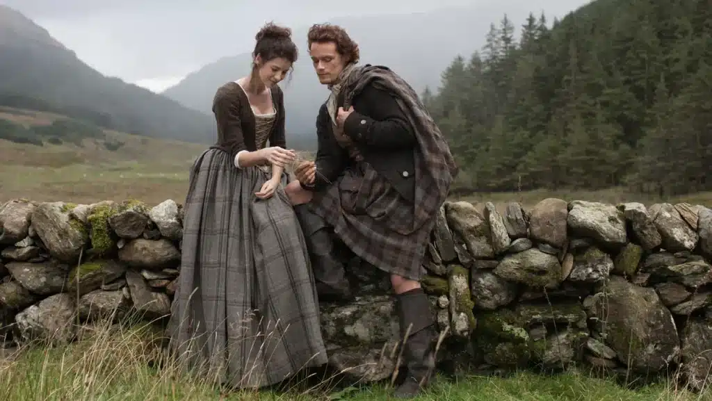 24 TV Series Set in Europe That'll Inspire Your Next Vacation 19 Outlander TV series promotional image with Claire and Jamie Fraser in 18th-century attire against a Scottish Highlands backdrop, representing historical TV shows set in Europe.