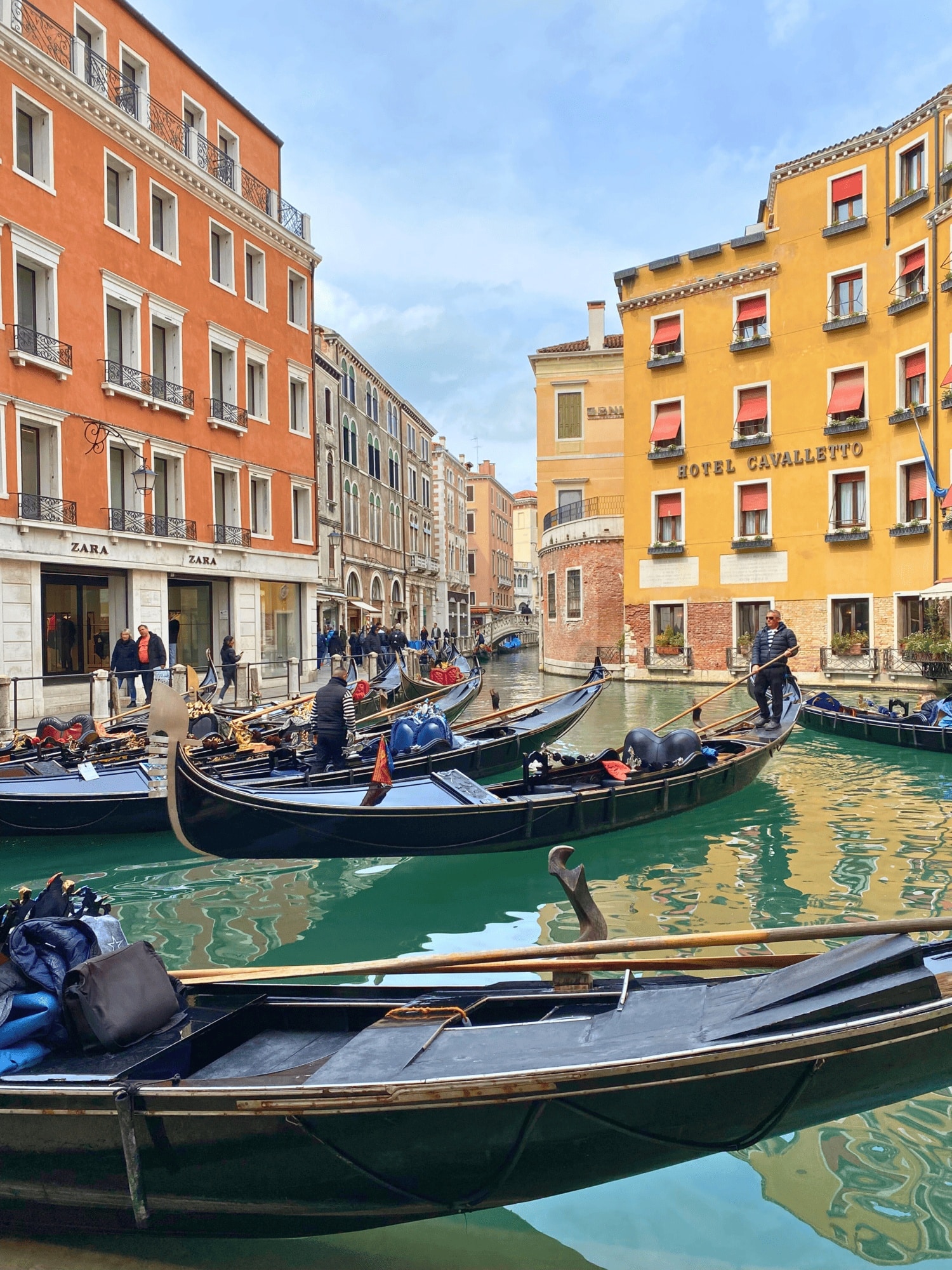 Italy 12 Classic Venetian gondolas gliding along a narrow canal in Venice, Italy, with striped‑shirt gondoliers steering the long black boats between sunlit buildings. The greenish water, pastel building facades, and shimmering reflections capture the romantic charm of a Venice, Italy gondola ride.